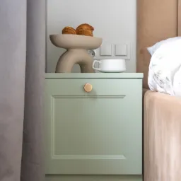 Nocny stolik w odcieniach zieleni z croissantami w dekoracyjnej misie i białą filiżanką, ustawiony obok ł&oacute;żka z beżowym zagł&oacute;wkiem i wzorzystą pościelą.