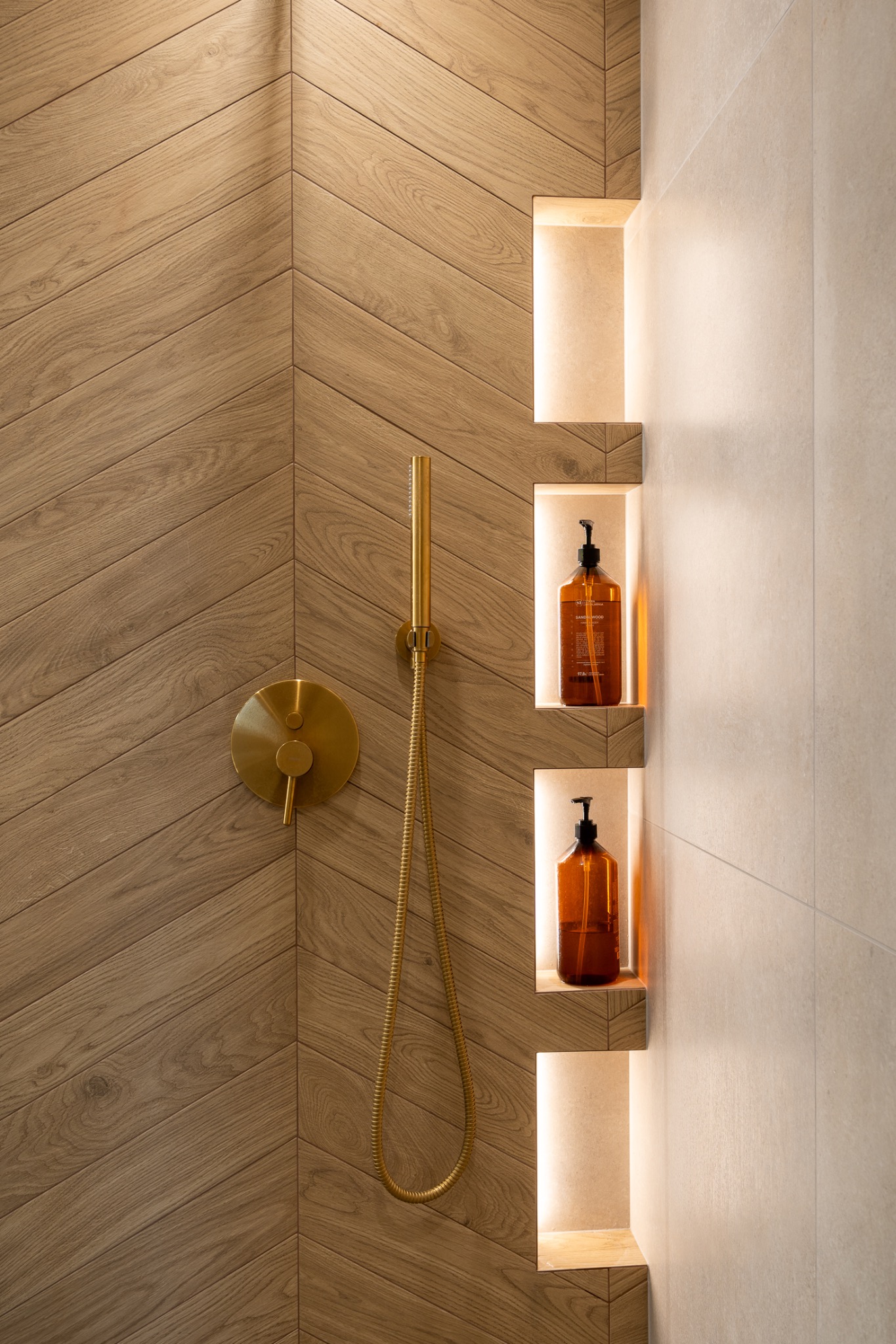 Nowoczesna łazienka z drewnianą ścianą ułożoną w jodełkę, złotą armaturą prysznicową i podświetlanymi niszami z brązowymi butelkami kosmetyków.