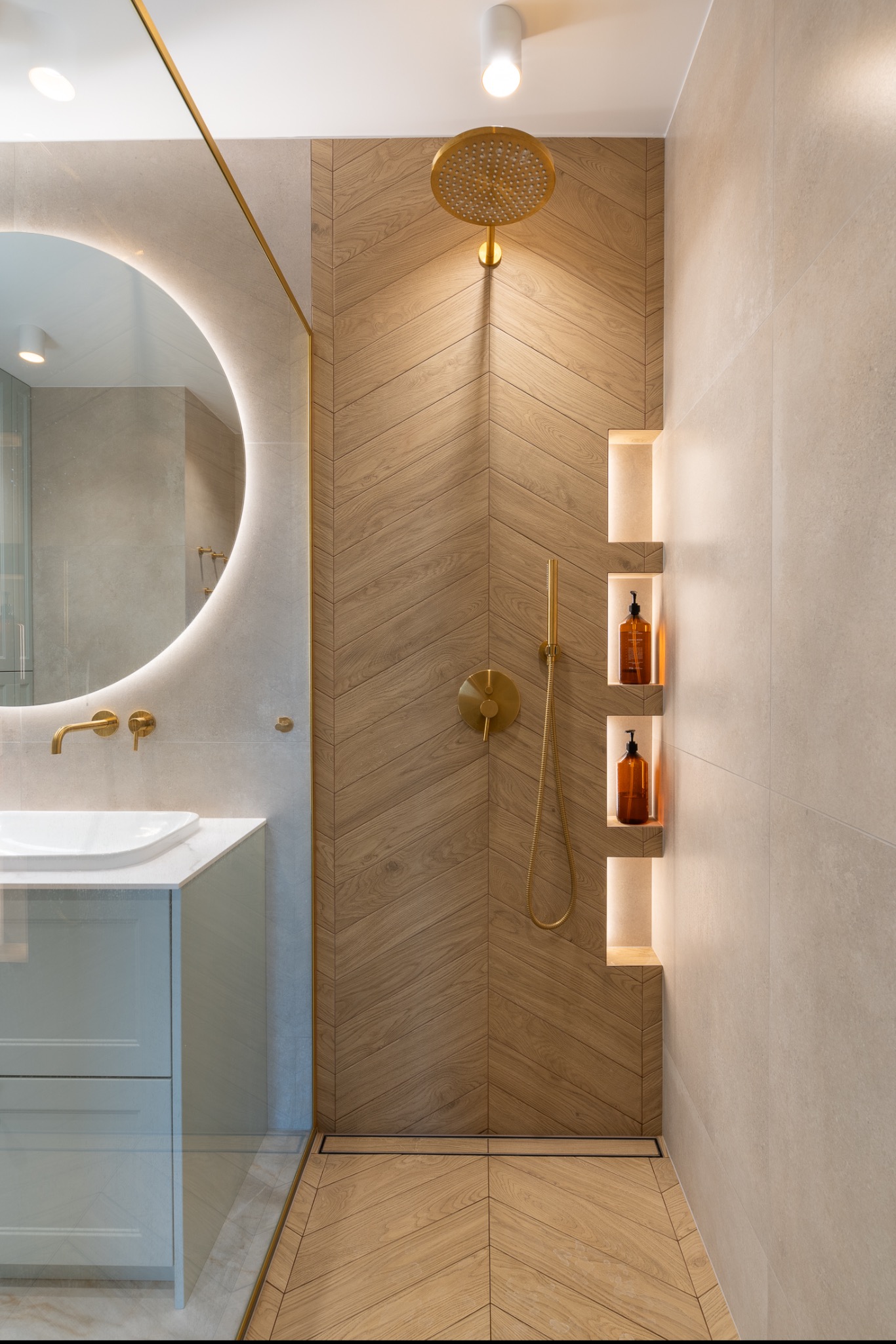 Nowoczesna łazienka z drewnopodobną ścianą prysznicową w jodełkę, złotą armaturą i okrągłym lustrem z podświetleniem LED.