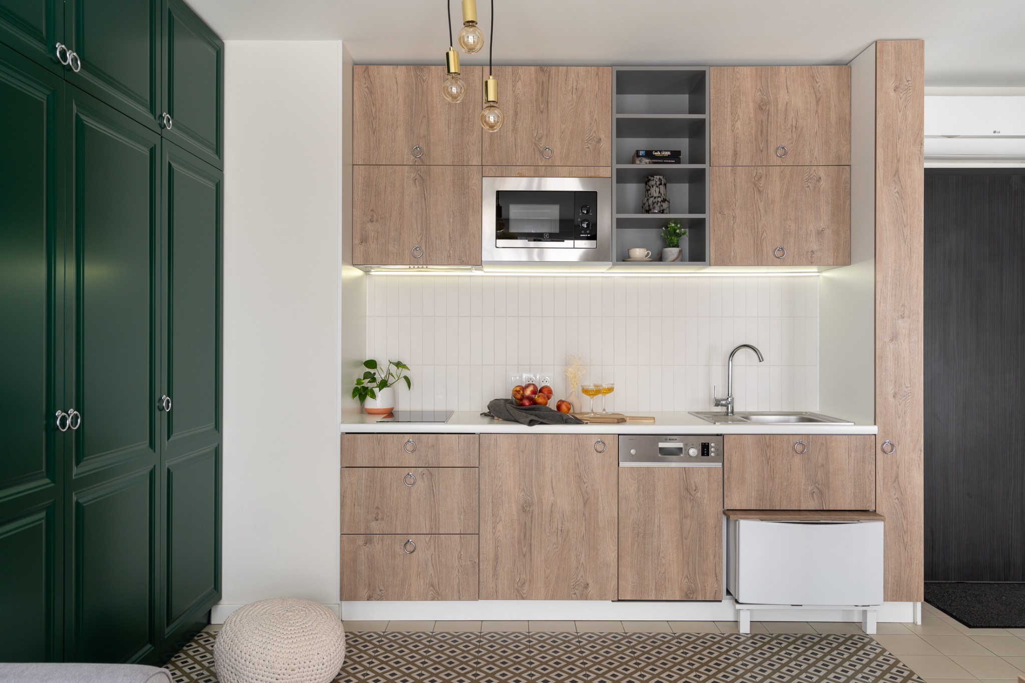 Nowoczesna kuchnia z drewnianymi frontami szafek, białymi płytkami na ścianie, zielonymi szafkami po lewej i wzorzystą podłogą. Na blacie widoczne owoce i szklanki z napojem.