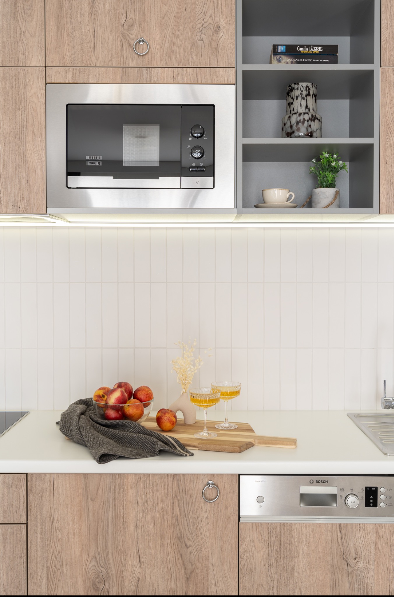 Jasna kuchnia z drewnianymi szafkami, mikrofalówką, zmywarką Bosch, owocami w szklanej misce, dwoma kieliszkami z żółtym napojem i drewnianą deską do krojenia na białym blacie.