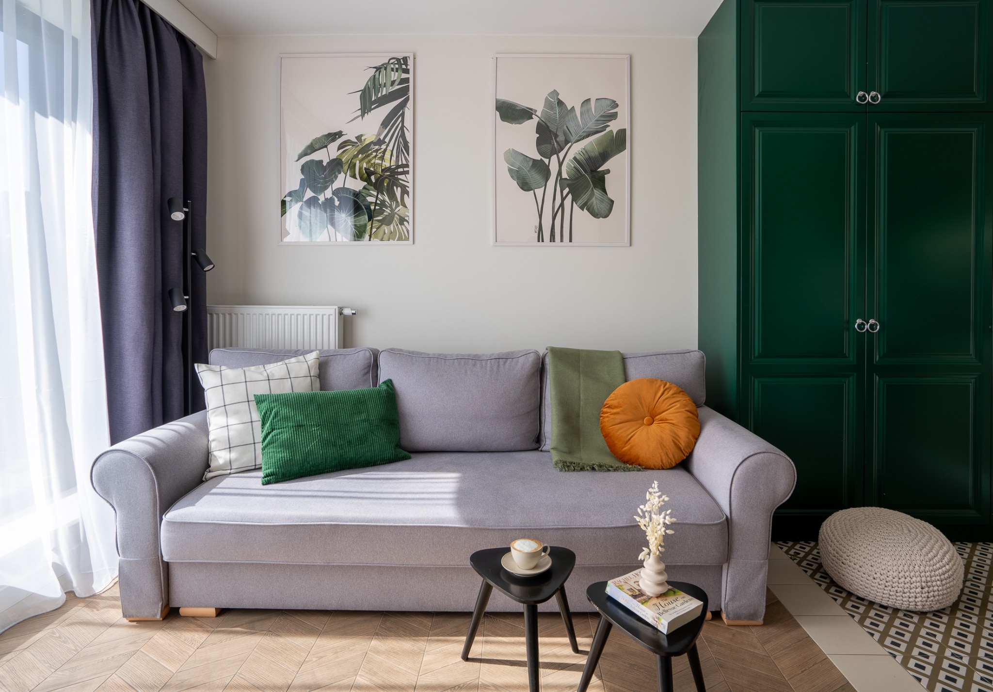 Jasnoszara sofa z zielonymi i wzorzystymi poduszkami, pomarańczową okrągłą poduszką i zielonym pledem, ustawiona na parkiecie w jodełkę, z dwoma czarnymi stolikami, na których stoi filiżanka kawy...