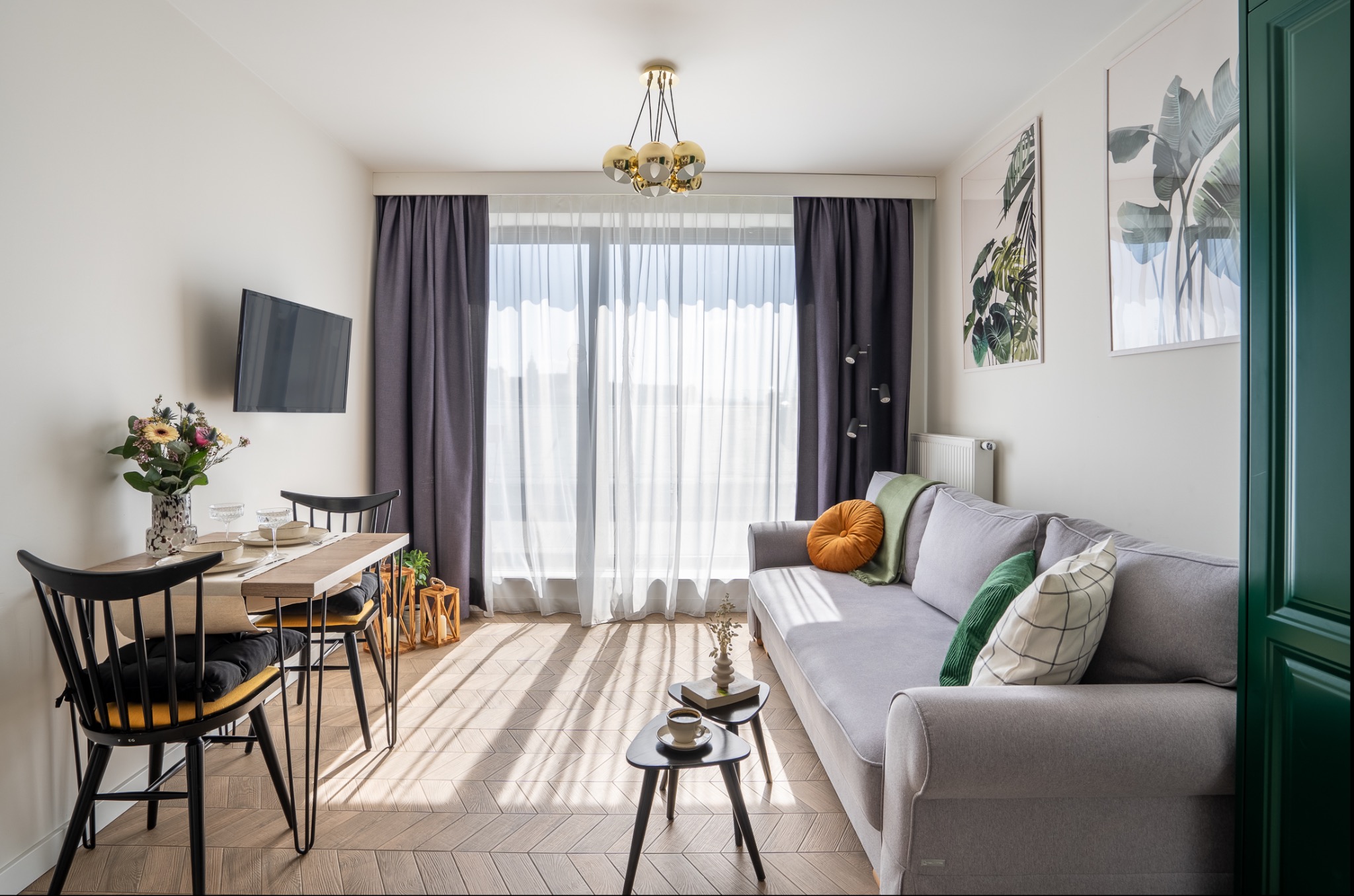 Jasne wnętrze mieszkania z szarą sofą z poduszkami, drewnianym stołem jadalnym z czarnymi krzesłami, telewizorem na ścianie i parkietem w jodełkę.