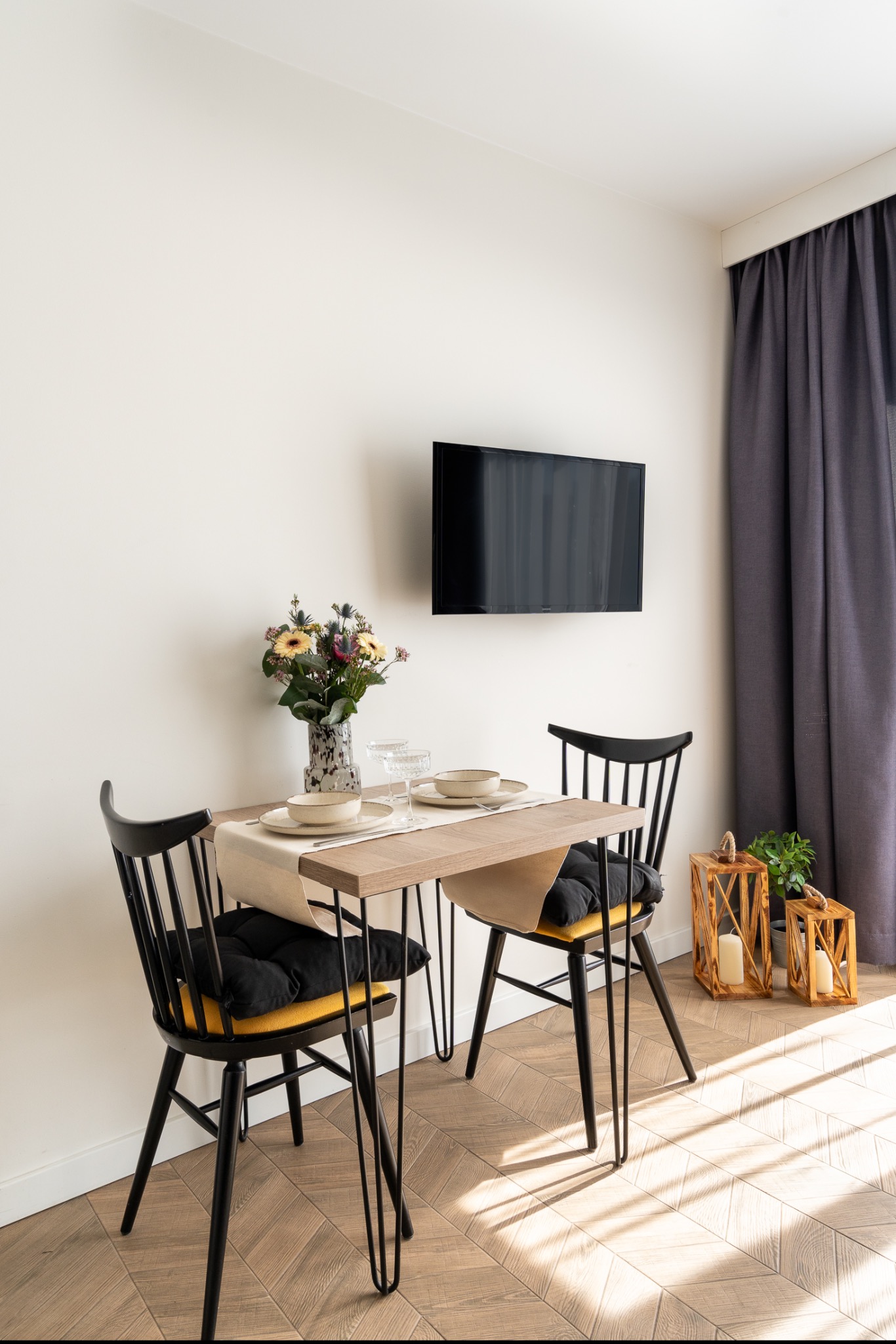 Jasne wnętrze jadalni z drewnianym stołem nakrytym dla dwóch osób, kwiaty w wazonie, czarne krzesła z poduszkami, telewizor na ścianie i drewniane lampiony ze świecami na podłodze.