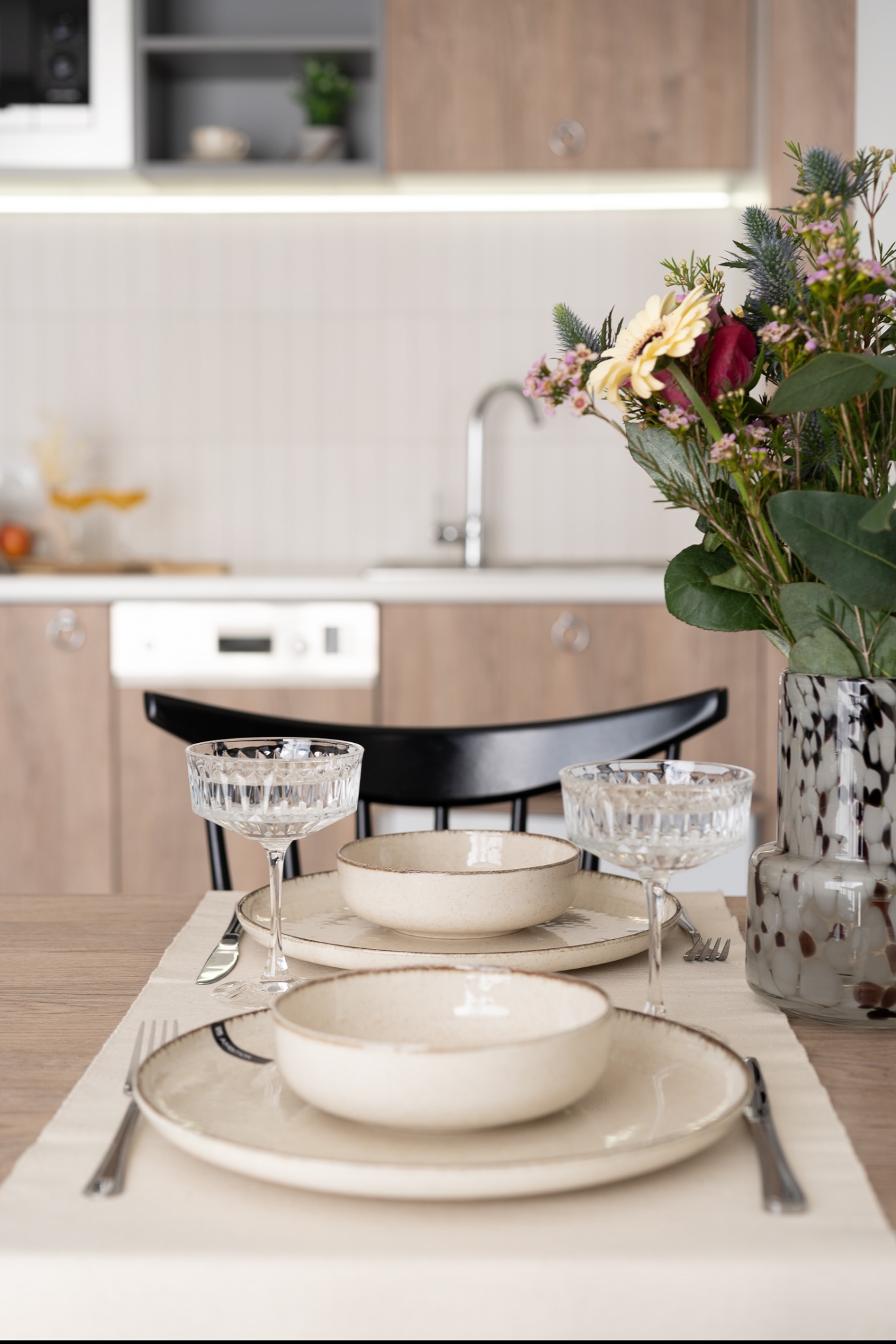 Ustawiony stół w jasnej kuchni z kwiatami w ozdobnym wazonie, kryształowymi kieliszkami i beżową zastawą na lnianym bieżniku.
