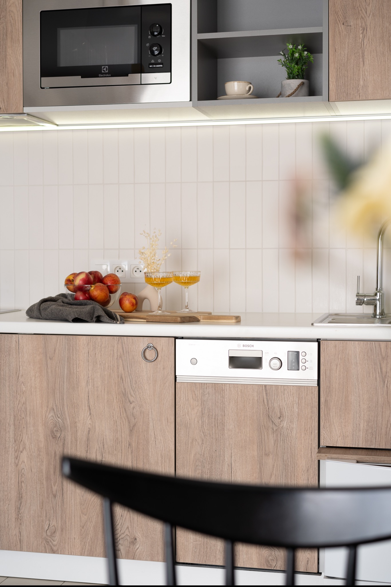 Jasna kuchnia z drewnianymi frontami, wyposażona w mikrofalówkę Electrolux, zmywarkę Bosch, z owocami w szklanej misie i dwoma kieliszkami z pomarańczowym napojem na blacie.