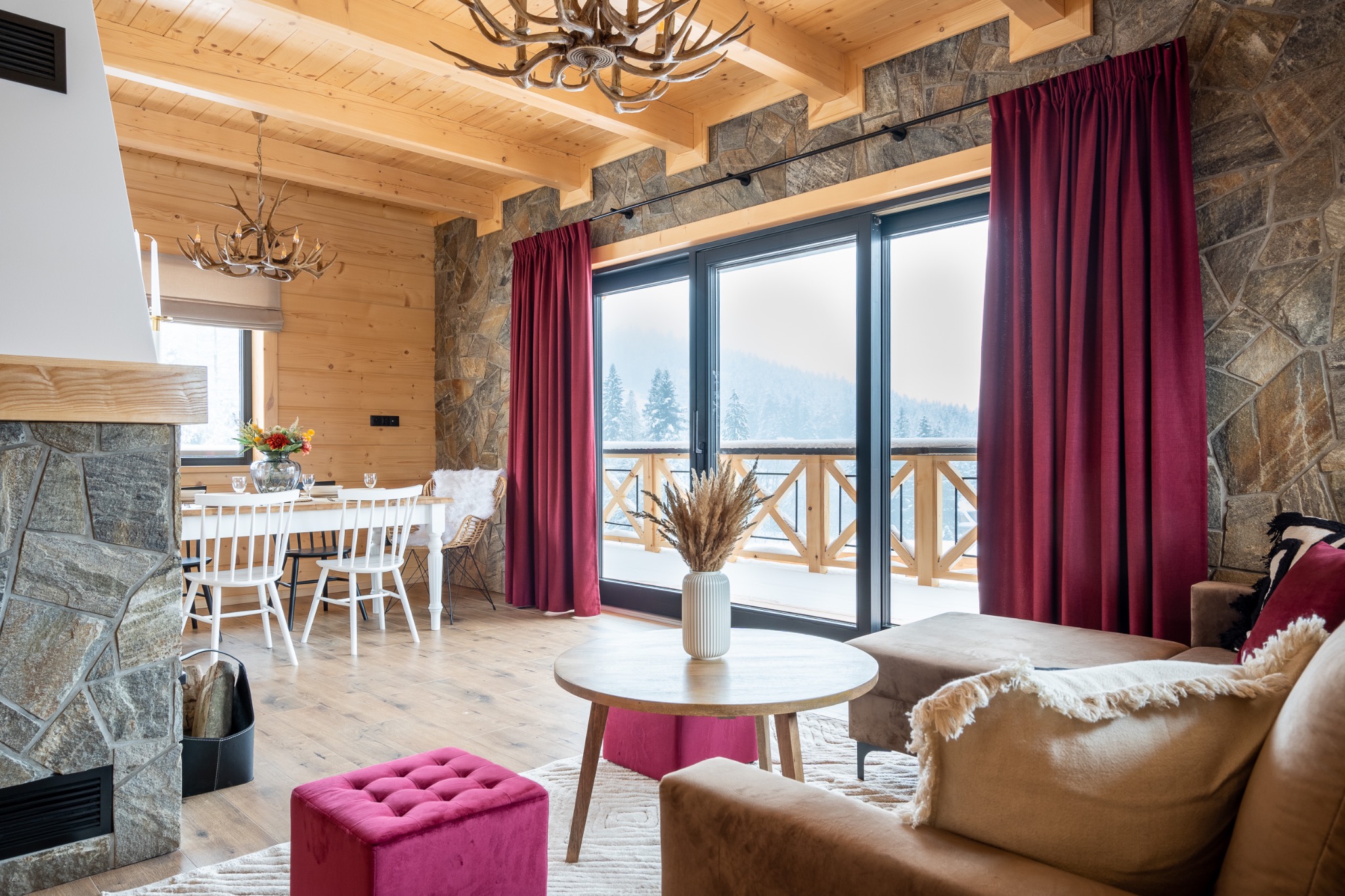 Rustykalny salon z drewnianymi ścianami, kamiennym kominkiem, sofą w odcieniach beżu i widokiem na ośnieżone góry przez duże okno z bordowymi zasłonami.