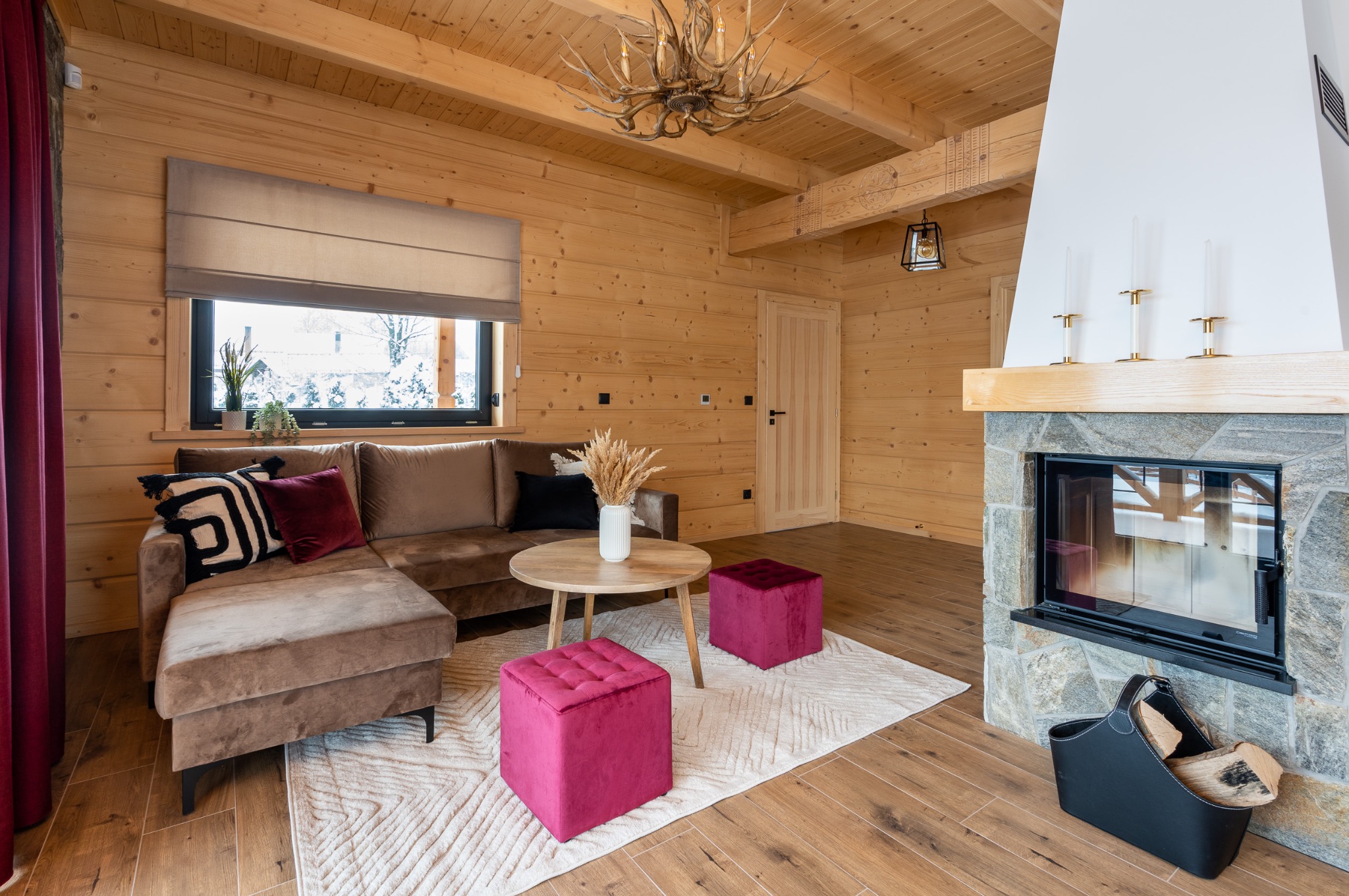 Wnętrze drewnianego domu z kominkiem, sofą w odcieniach brązu, okrągłym stolikiem, dwoma pufami w kolorze fuksji i dekoracyjnym żyrandolem z poroża.