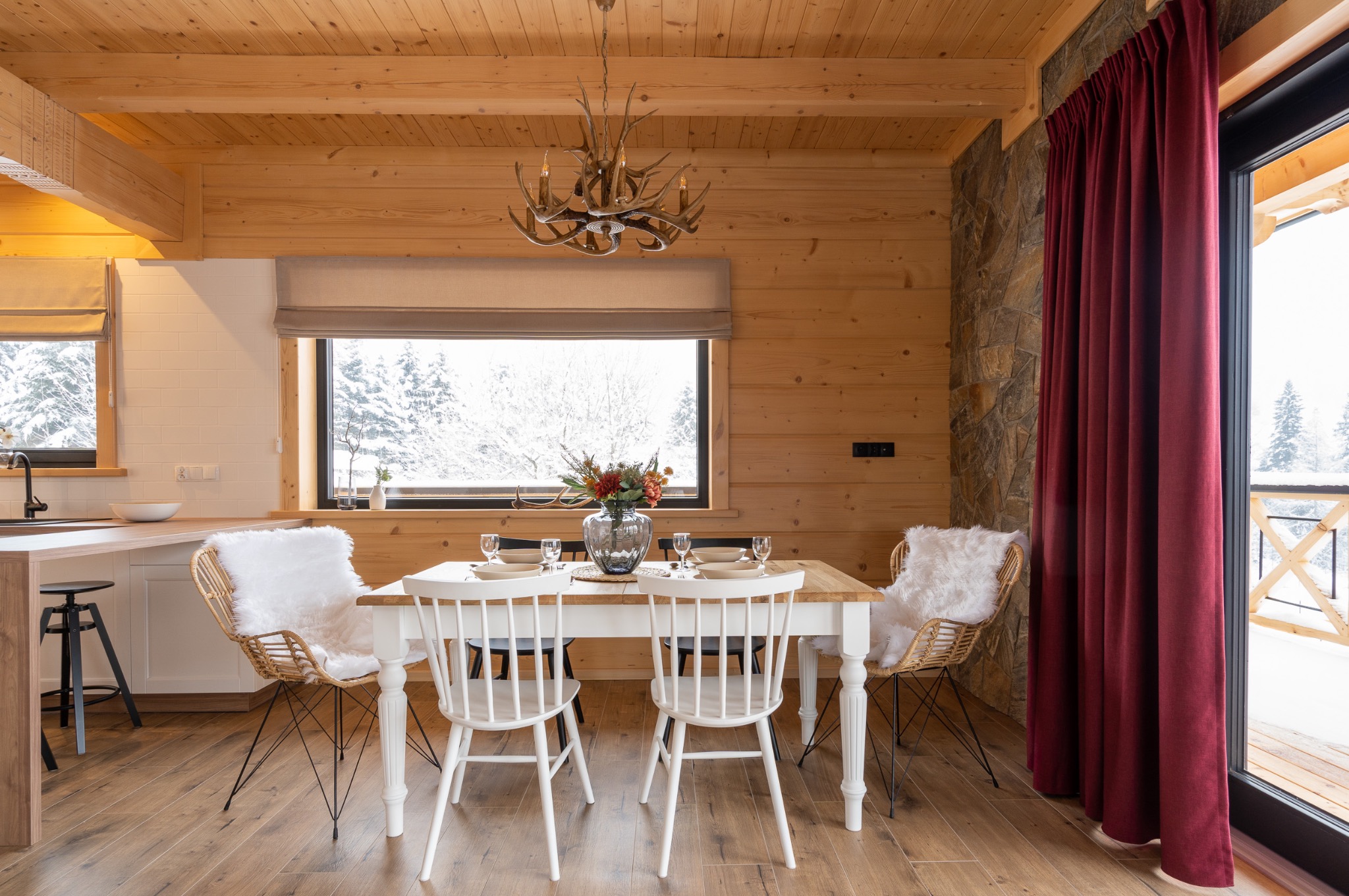 Drewniana jadalnia w góralskim stylu z białym stołem nakrytym dla czterech osób, widok na zaśnieżony krajobraz przez okno, oryginalny żyrandol z poroża.