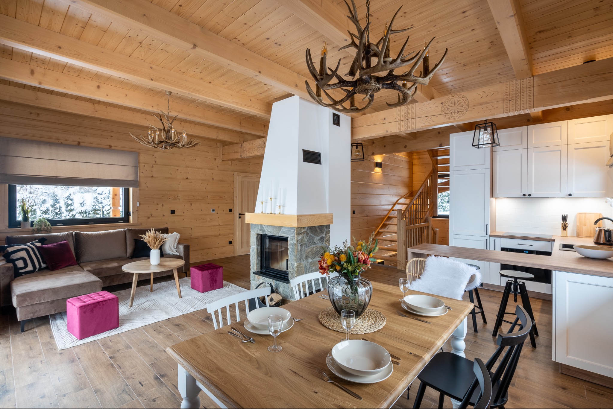 Jasne, drewniane wnętrze domu z kominkiem, stołem jadalnym z zastawą, sofą i kuchnią w tle, oświetlone lampą z poroża.