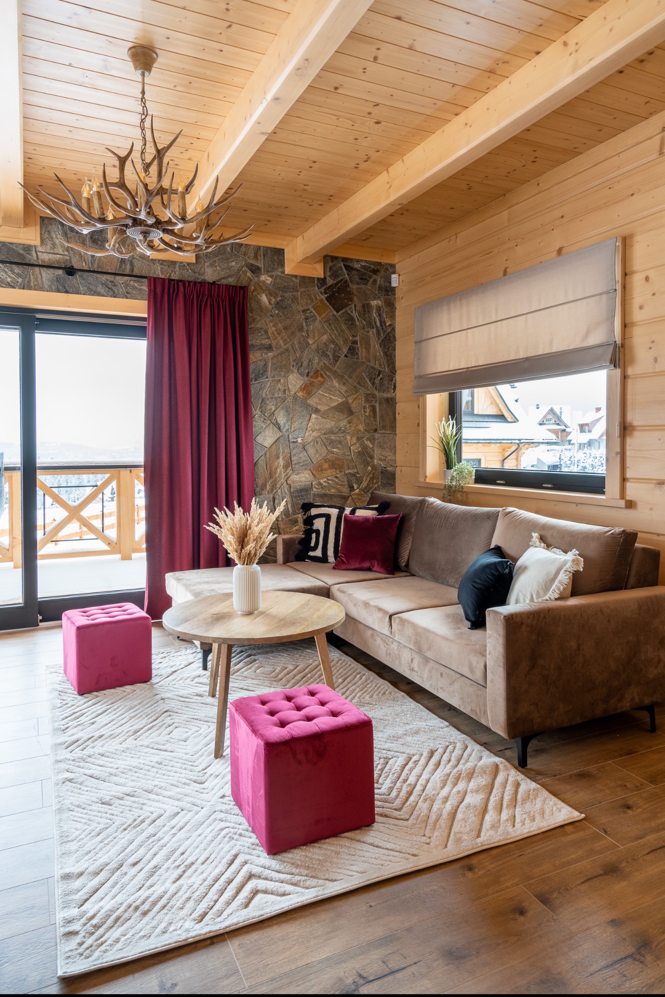 Jasne drewniane wnętrze góralskiego salonu z beżową sofą narożną, okrągłym stolikiem kawowym, dwoma różowymi pufami i żyrandolem z poroża.
