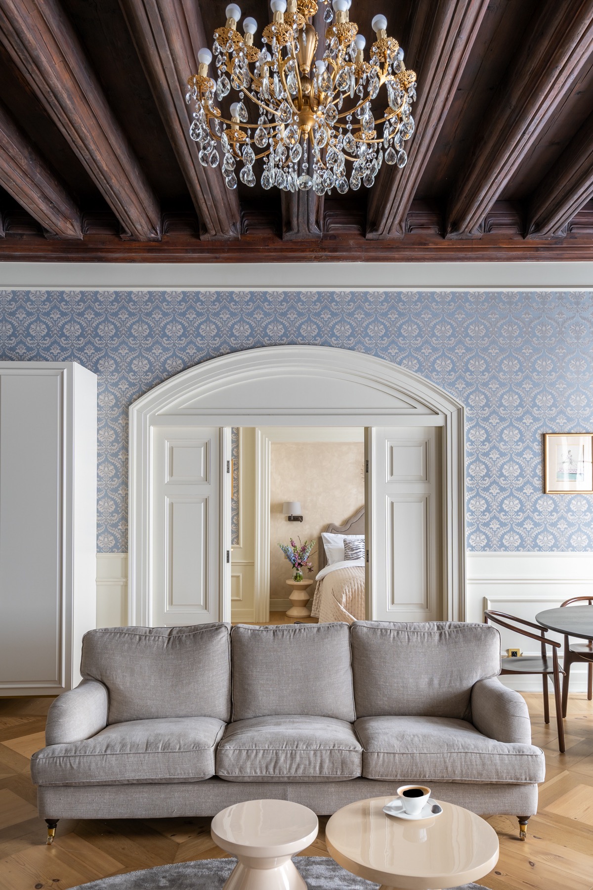 Eleganckie wnętrze salonu z szarą sofą, okrągłymi stolikami kawowymi, kryształowym żyrandolem i widokiem na sypialnię przez łukowe drzwi.