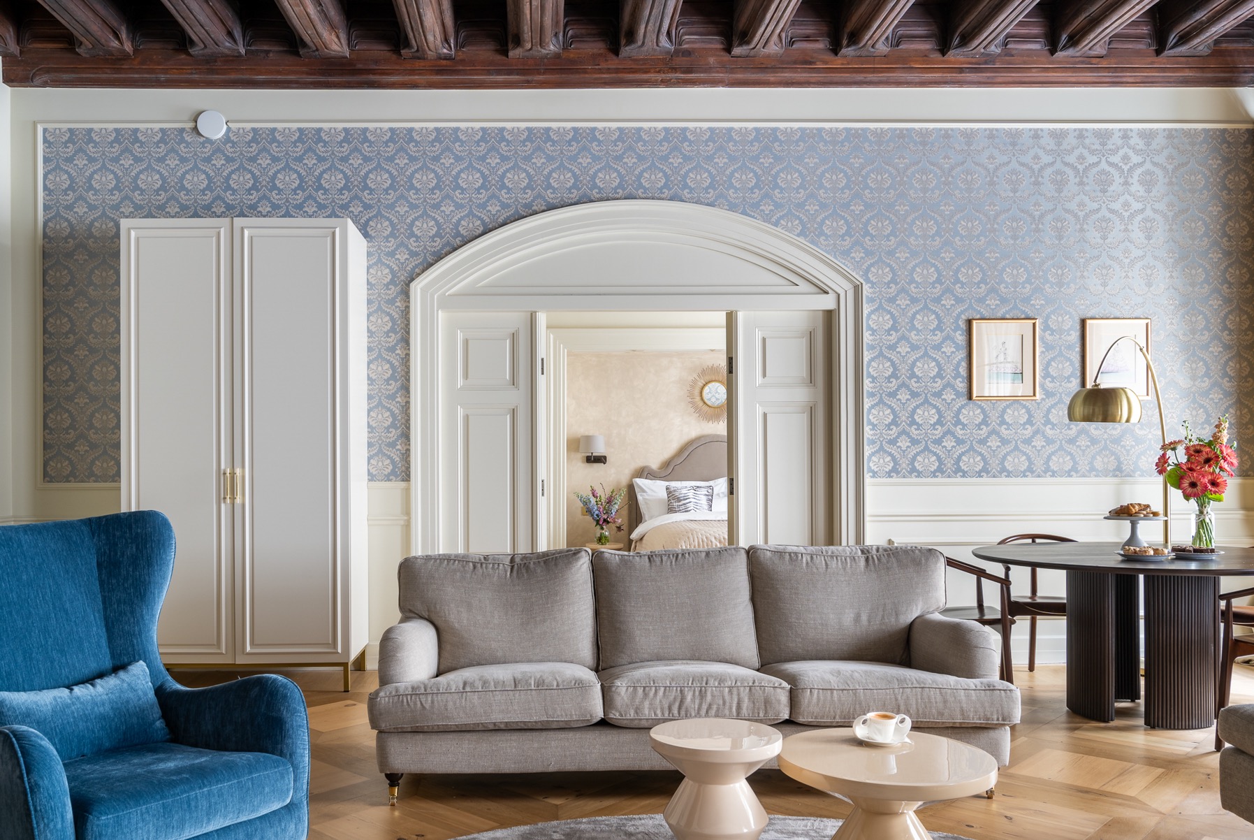 Eleganckie wnętrze apartamentu z szarą sofą, błękitnymi wzorzystymi tapetami, drewnianym sufitem i widokiem na sypialnię z okrągłym lustrem.