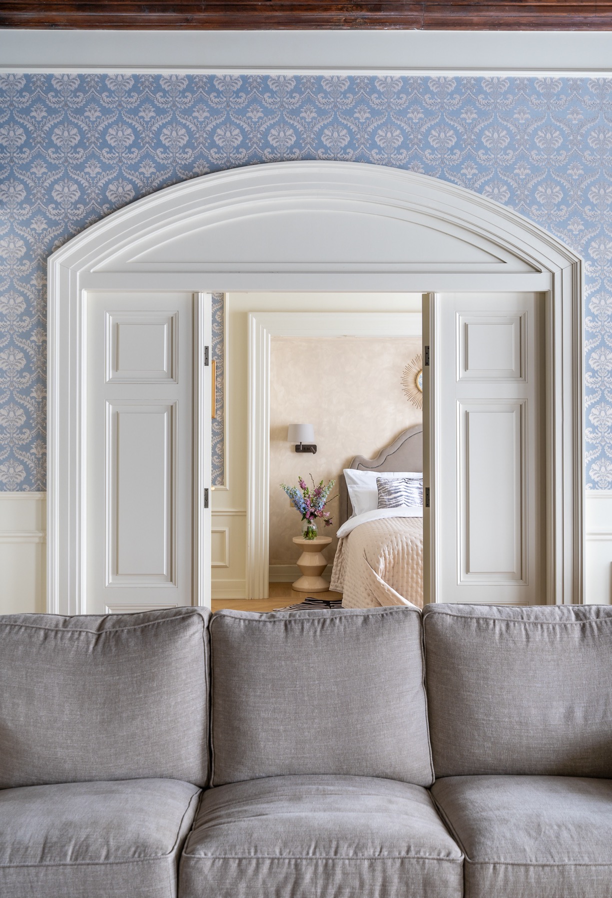 Eleganckie wnętrze sypialni widziane zza szarej sofy, przez białe, przesuwne drzwi z łukowym nadprożem, na ścianach błękitna tapeta w delikatny wzór, w sypialni kwiaty w wazonie na stoliku nocnym.