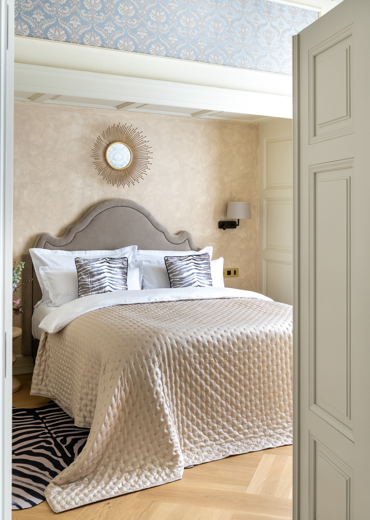 Elegancka sypialnia w odcieniach beżu z łóżkiem z pikowaną narzutą, poduszkami w zebrę, okrągłym lustrem w kształcie słońca i wzorzystą tapetą na suficie, widziana zza uchylonych drzwi.