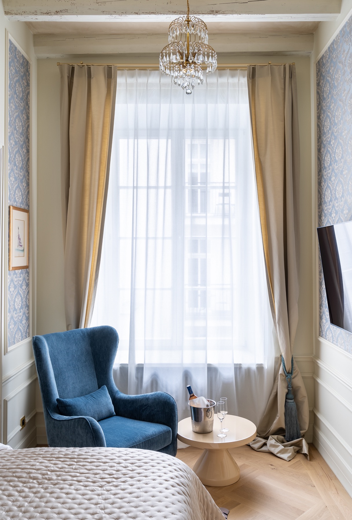 Elegancki pokój hotelowy z niebieskim fotelem, stolikiem z szampanem w wiaderku z lodem i dwoma kieliszkami, zasłoniętym oknem i kryształowym żyrandolem.