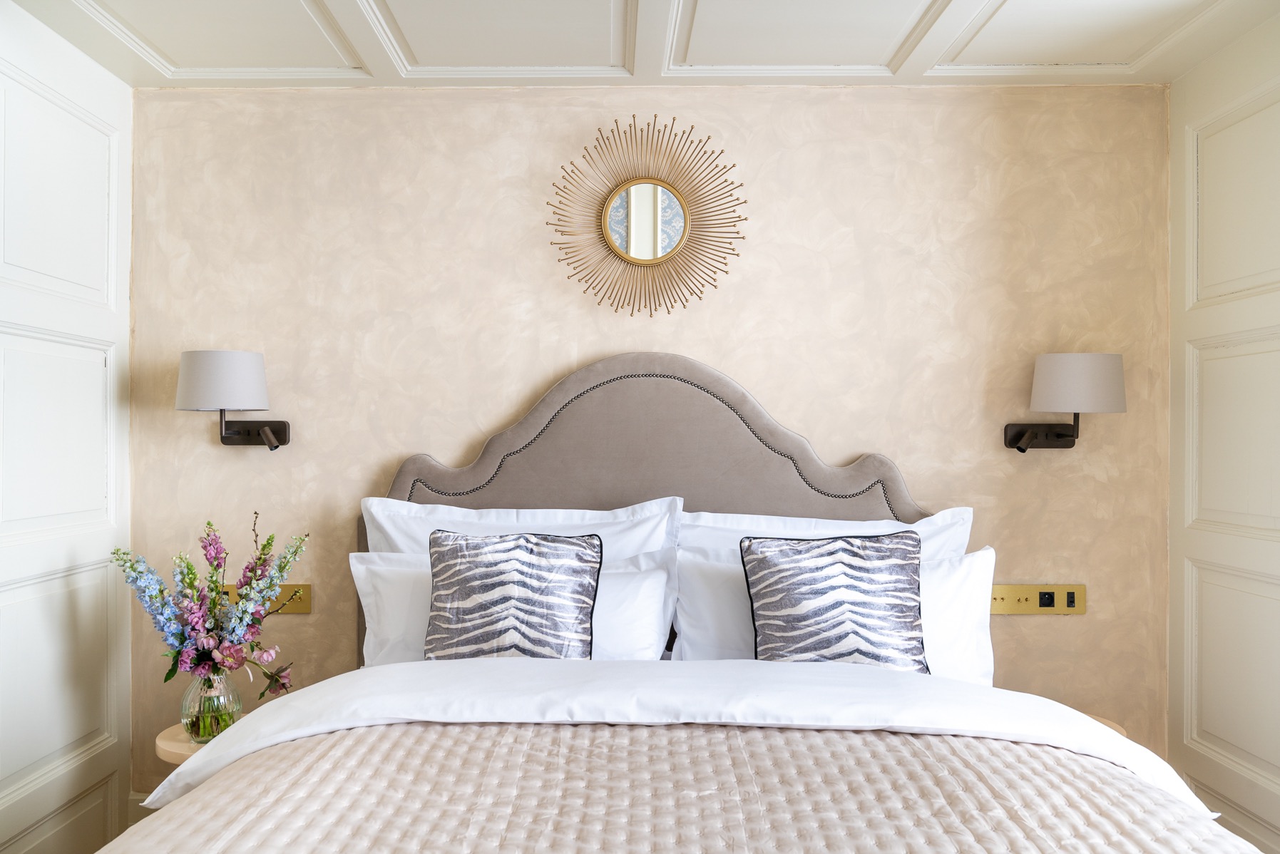 Elegancka sypialnia z beżowymi ścianami, dekoracyjnym lustrem w kształcie słońca nad wezgłowiem łóżka, kwiatami w wazonie na stoliku nocnym i lampkami ściennymi.