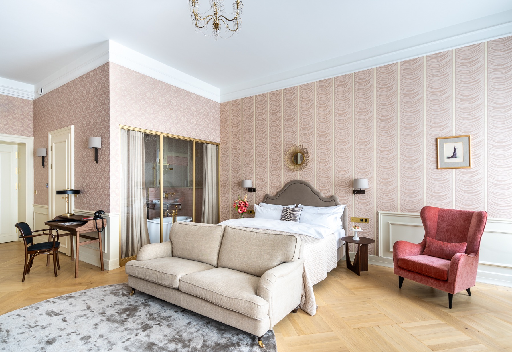 Elegancki pokój hotelowy z łazienką za szklaną ścianą, beżowa sofa na dywanie, łóżko z szarą ramą, czerwony fotel uszak i biurko z krzesłem w stylu retro.