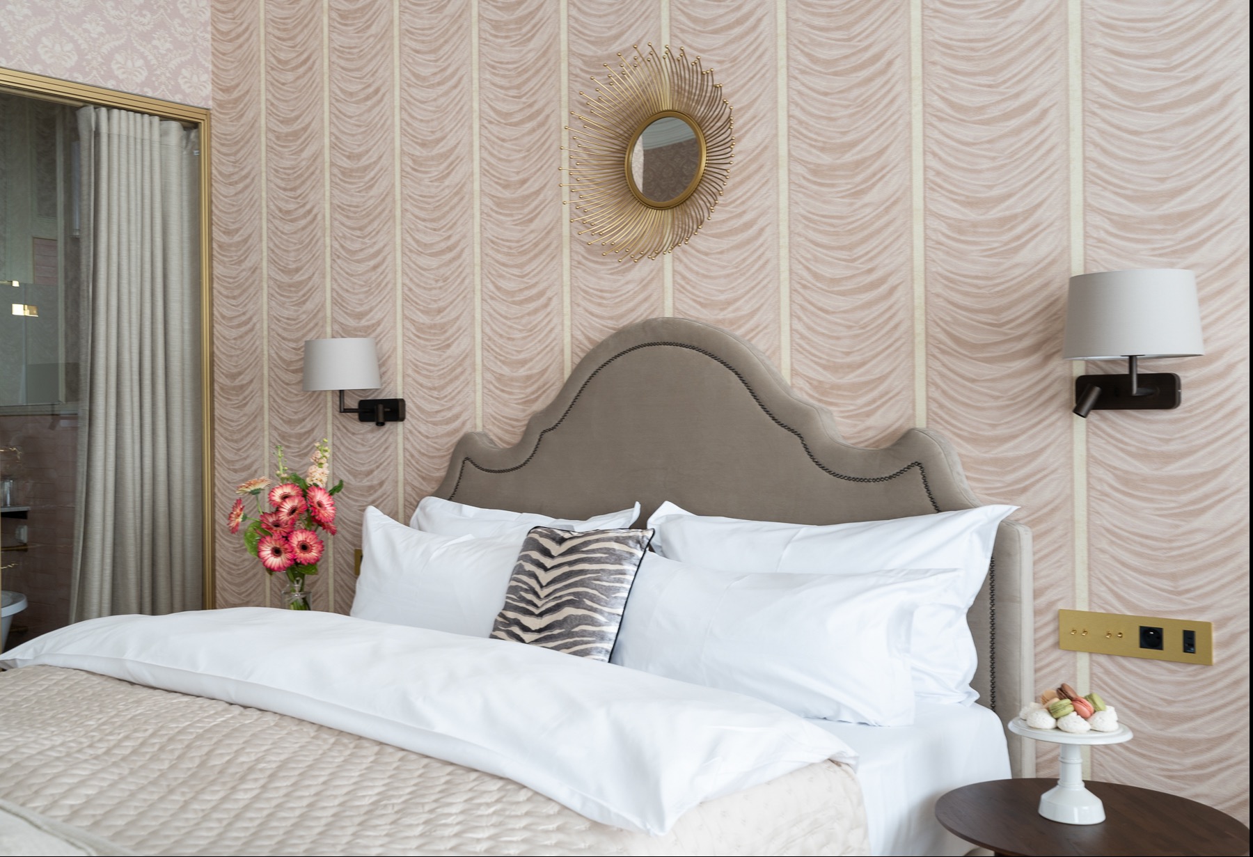 Elegancka sypialnia z łóżkiem z szarym zagłówkiem, białymi poduszkami i narzutą w kolorze beżowym, na stoliku nocnym talerz z makaronikami, a na ścianie lustro w złotej ramie w kształcie słońca.