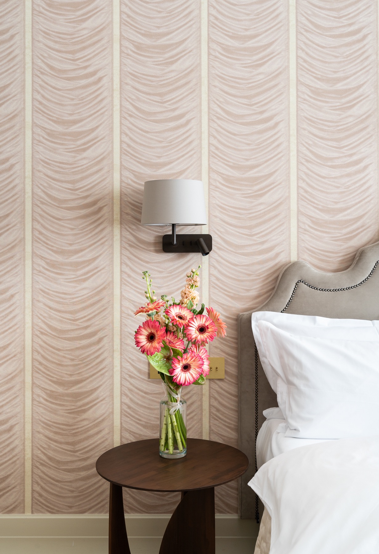 Bukiet różowych gerber w szklanym wazonie na okrągłym, drewnianym stoliku nocnym, obok łóżka z szarym zagłówkiem i białą pościelą, na tle ściany w beżowe pasy i lampki nocnej.