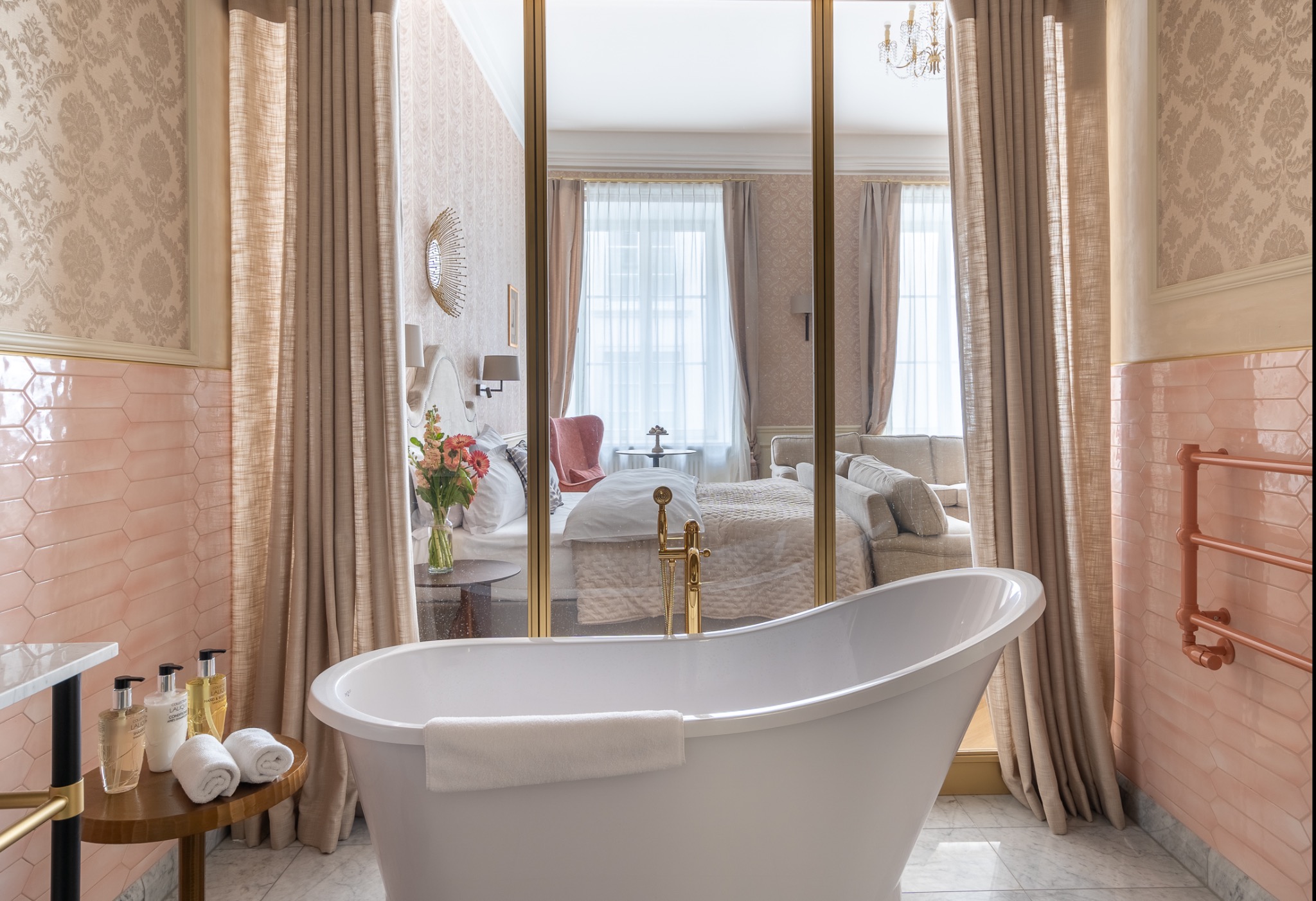 Elegancka łazienka z wolnostojącą wanną, widok na sypialnię przez szklaną ścianę, pastelowe płytki i marmurowa podłoga, aranżacja wnętrza w Krakowie.