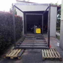 Metalowa konstrukcja szkieletowa na paletach przed garażem, w tle żółta spawarka, widoczne spawy.