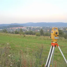 Żółty teodolit ustawiony na trójnogu na łące z widokiem na miasto w oddali, wczesny poranek.