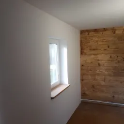 Pokój po remoncie: białe ściany, drewniana boazeria na jednej ścianie, drewniany parapet, widoczne kable w rogu.