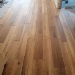 Ułożone panele podłogowe w odcieniach brązu, widoczne ślady po pracach wykończeniowych i odkurzacz przemysłowy w rogu pomieszczenia.