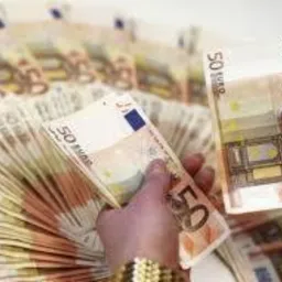 Dłonie trzymające wachlarz banknotów 50 euro, ostrość na banknot w prawej dłoni, w tle rozmyty stos banknotów.