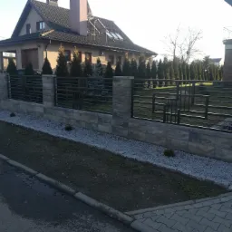 Nowoczesne ogrodzenie z metalowymi przęsłami i betonową podmurówką, ozdobione drobnym kamieniem i niskimi krzewami, na tle domu z brązową elewacją i rzędem iglaków.