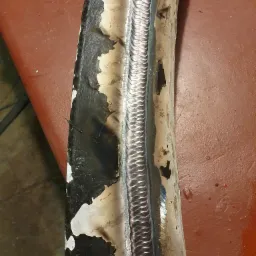 Spoina na zakrzywionej metalowej rurze z widocznymi śladami korozji i resztkami farby, umieszczona na czerwonym podłożu. Ukazanie detali i precyzji wykonania.