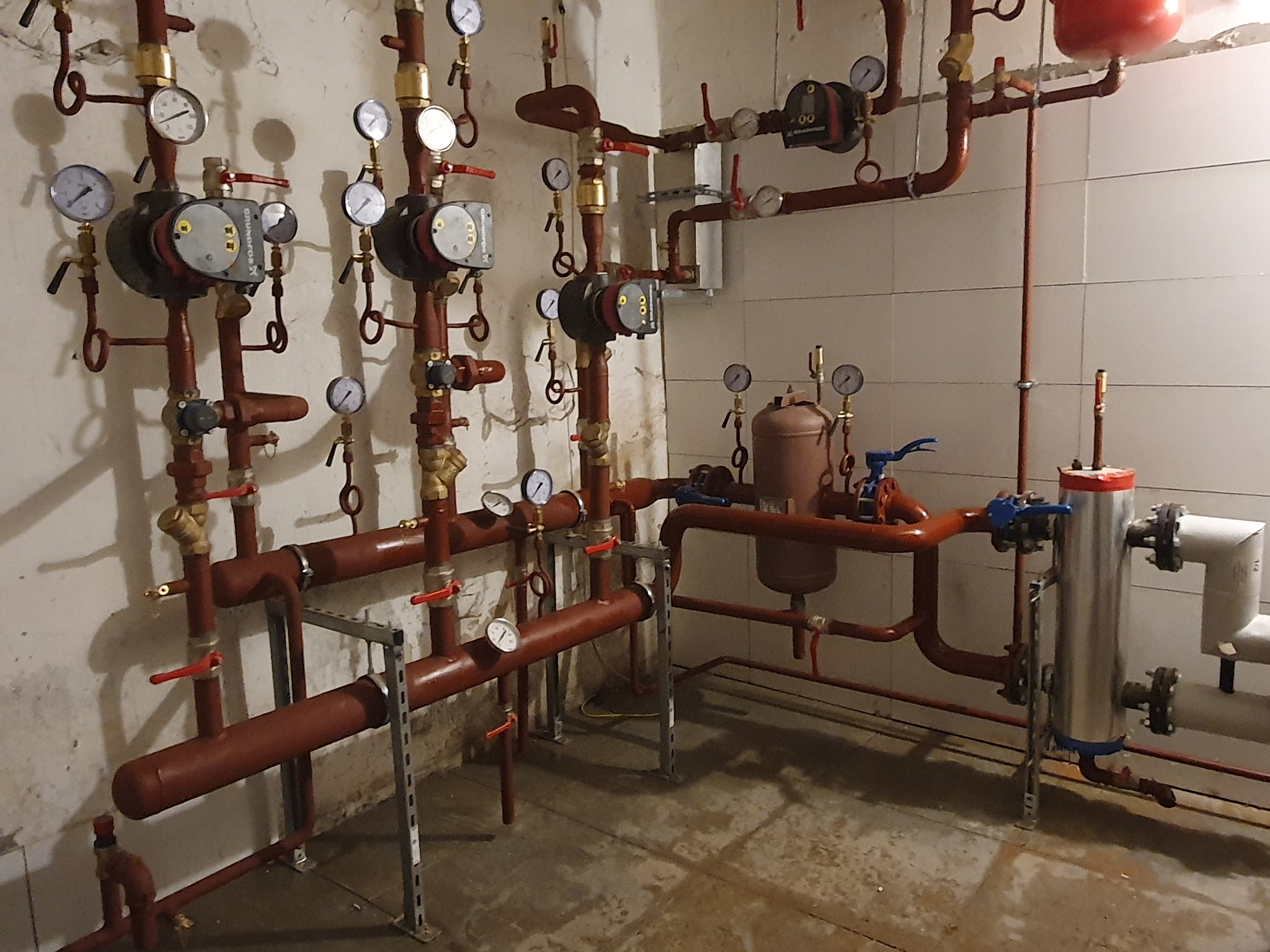 Złożona instalacja grzewcza z czerwonymi rurami, manometrami i pompami obiegowymi na tle białych płytek. Widoczne spawane połączenia i elementy mocujące.