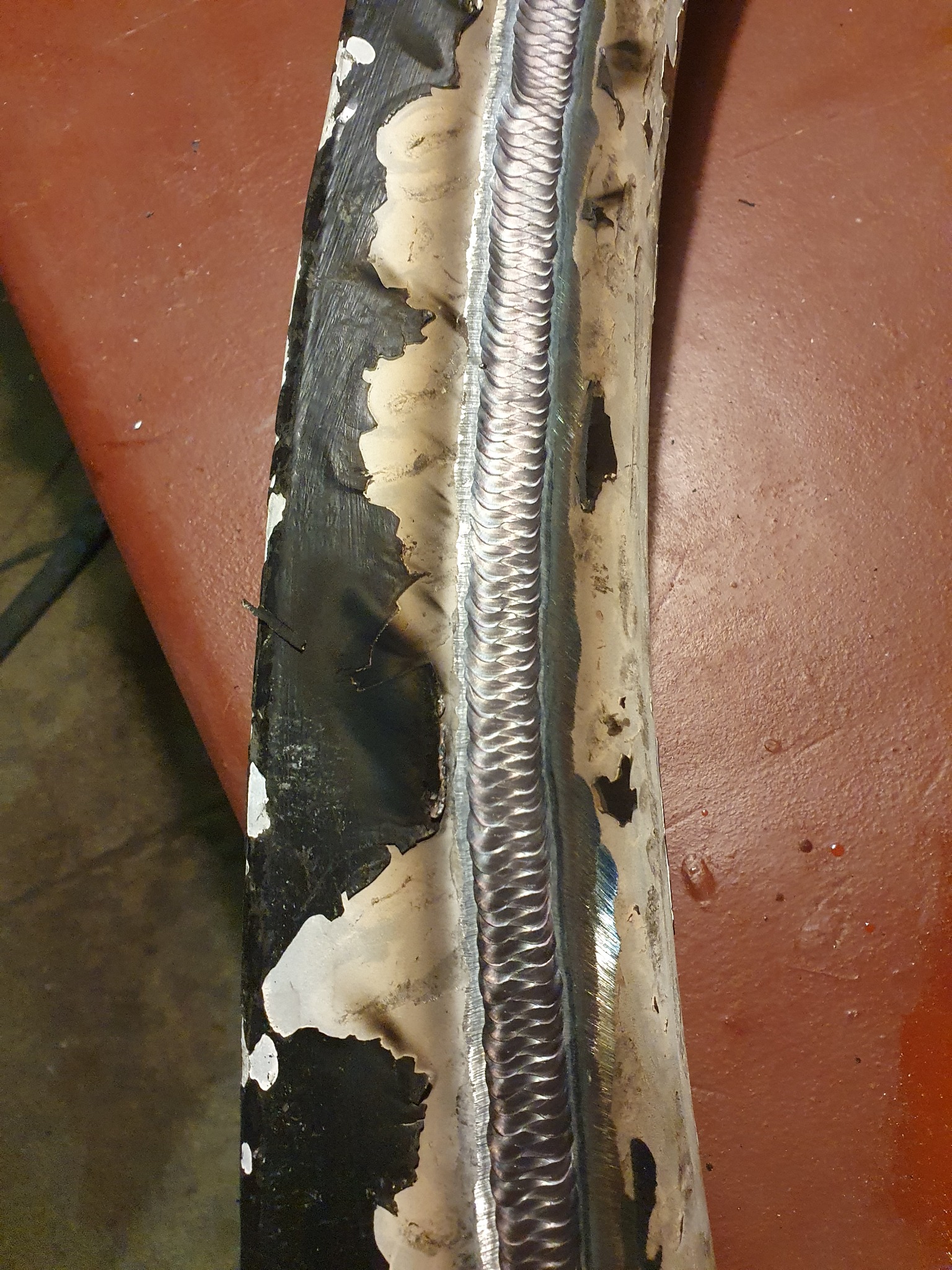 Spoina na zakrzywionej metalowej rurze z widocznymi śladami korozji i resztkami farby, umieszczona na czerwonym podłożu. Ukazanie detali i precyzji wykonania.