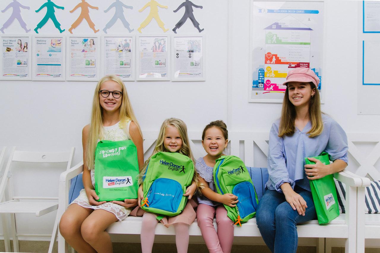 Grupa uśmiechniętych dzieci i młoda kobieta siedzą na białej ławce, trzymając zielone torby i plecaki z logo Helen Doron English. Na ścianie wiszą plakaty z informacjami o kursach językowych...