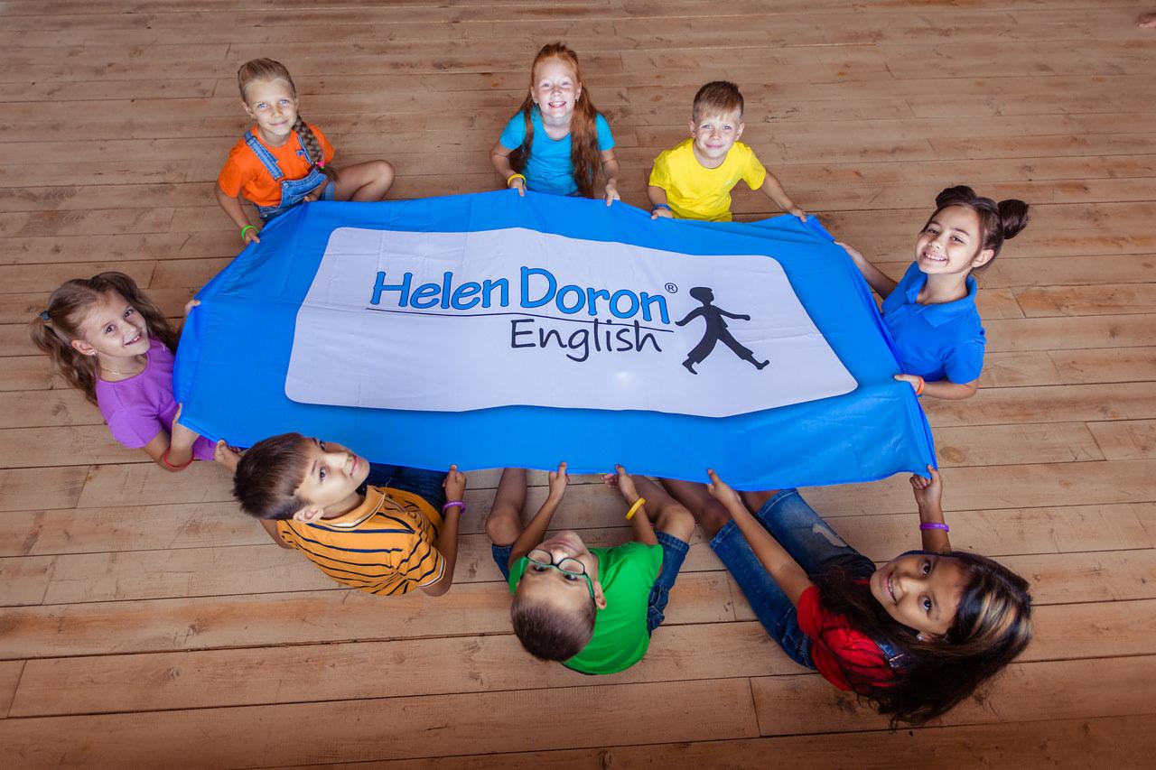 Grupa uśmiechniętych dzieci trzymająca baner z napisem 'Helen Doron English' na drewnianej podłodze, widok z góry.