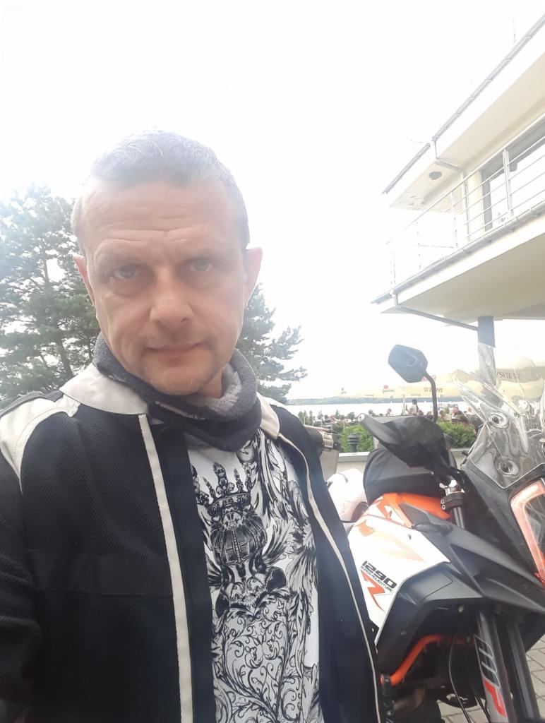 Mężczyzna w kurtce motocyklowej pozuje obok pomarańczowo-białego motocykla 1290 w Warszawie, w tle widoczny budynek i drzewa.
