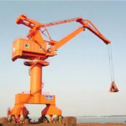 Pomarańczowy dźwig portowy z kabiną i wysięgnikiem, w tle błękitne niebo i fragment nabrzeża. Widoczny mechanizm podnoszenia z ładunkiem.