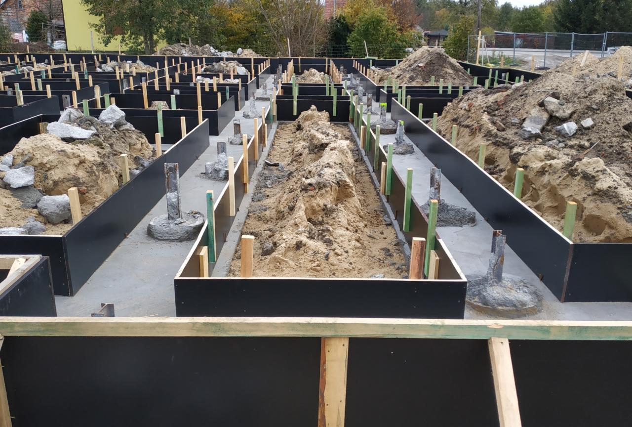 Szeroki kadr na przygotowany szalunek pod wylewkę fundamentów z widocznymi wystającymi prętami zbrojeniowymi i nasypaną ziemią wewnątrz niektórych sekcji.