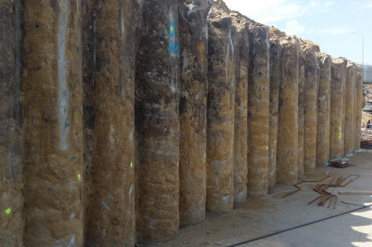 Zabezpieczenie wykopu palisadą z pali wierconych, widoczne oznaczenia geodezyjne na palach i zbrojenie na ziemi.