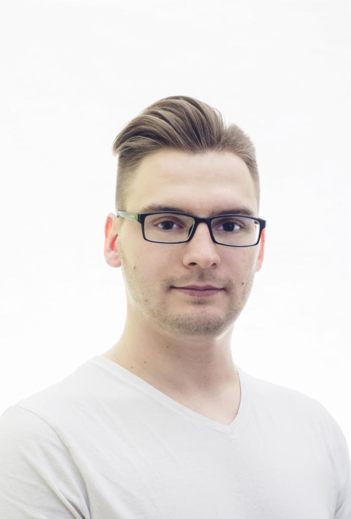 Portret młodego mężczyzny w okularach na białym tle. Krótkie włosy zaczesane do góry, biała koszulka z dekoltem w serek. Ujęcie od klatki piersiowej w górę.