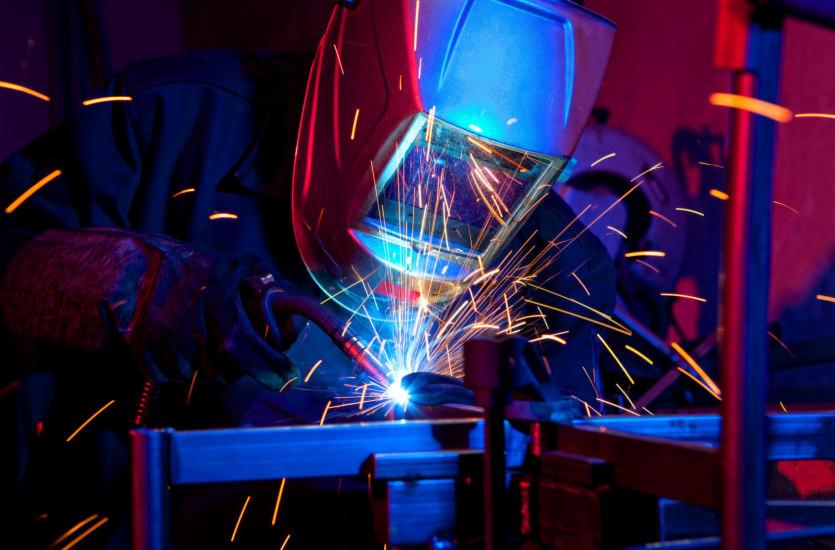 Pracownik w masce ochronnej łączy metalowe elementy, w tle widoczne iskry i ciemne otoczenie. Blask spawania oświetla stanowisko pracy.
