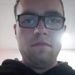 Portret mężczyzny w okularach z czarną bluzą, ujęcie z dołu. Widoczne odbicia w szkłach okularów. Twarz oświetlona z góry, krótko ostrzyżone włosy.