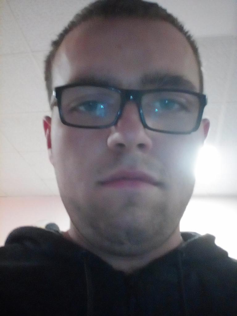 Portret mężczyzny w okularach z czarną bluzą, ujęcie z dołu. Widoczne odbicia w szkłach okularów. Twarz oświetlona z góry, krótko ostrzyżone włosy.