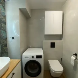 Nowoczesna łazienka z drewnianą podłogą, umywalką nablatową, pralką Bosch i wiszącą toaletą. Na ścianach szare płytki i tapeta w liście.