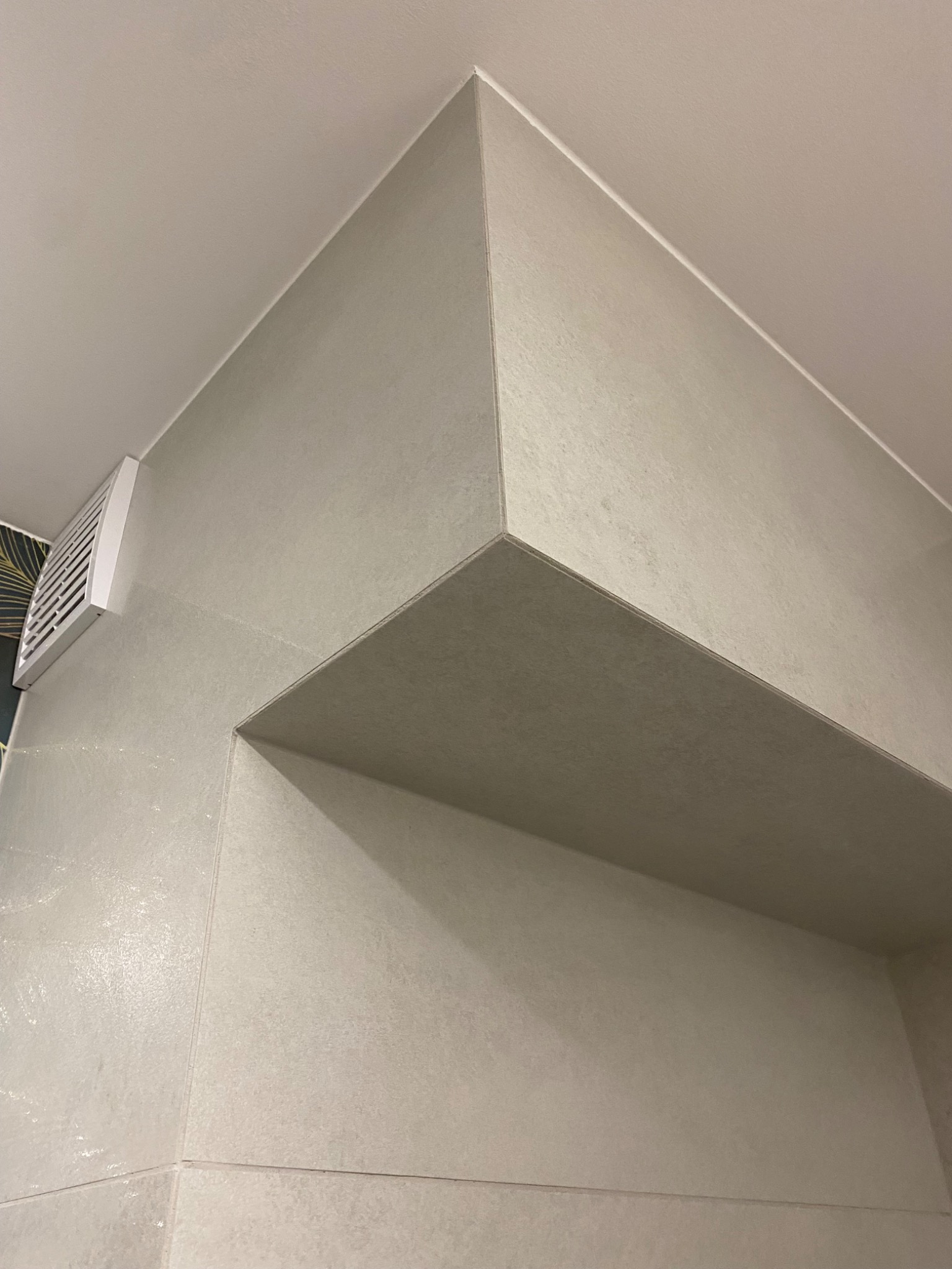 Wykończona łazienka z jasnymi płytkami imitującymi kamień, widoczny narożnik z półką i kratka wentylacyjna.