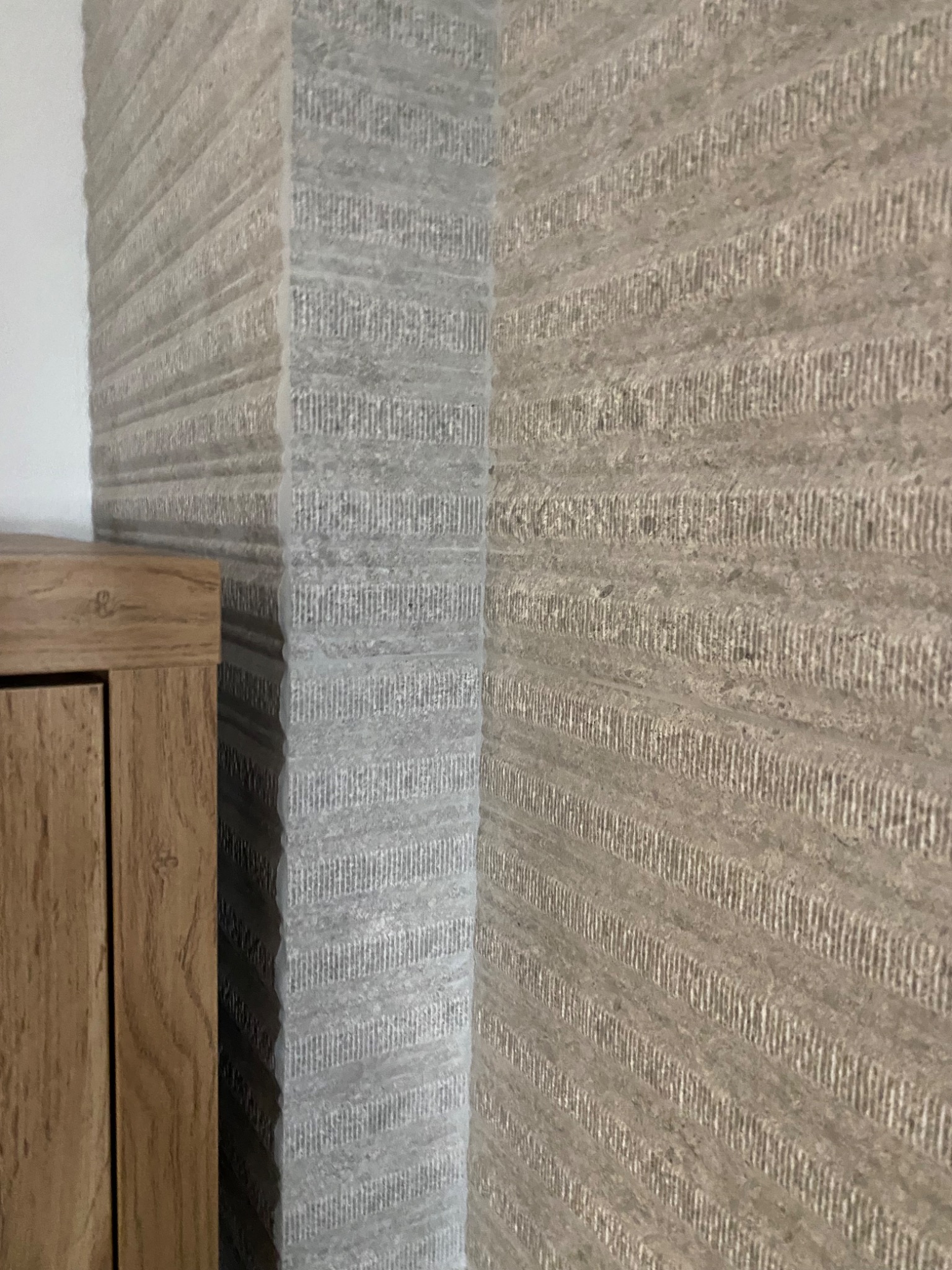 Strukturalna ściana z szarego kamienia z poziomymi rowkami, narożnik z listwą i fragment drewnianego mebla.