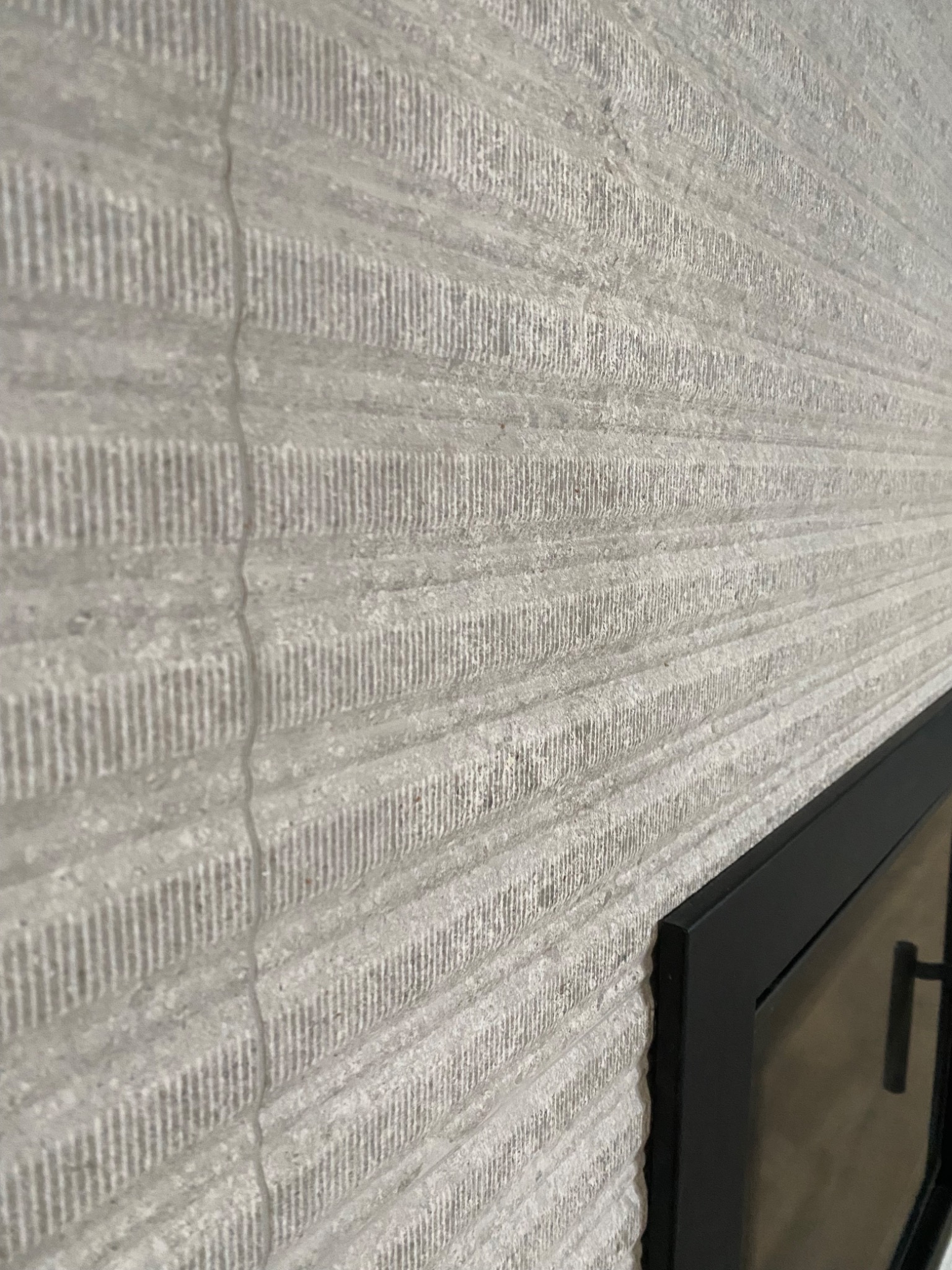 Szara ściana z dekoracyjną fakturą przypominającą poziome pasy, przylegająca do czarnej ramy, prawdopodobnie kominka.