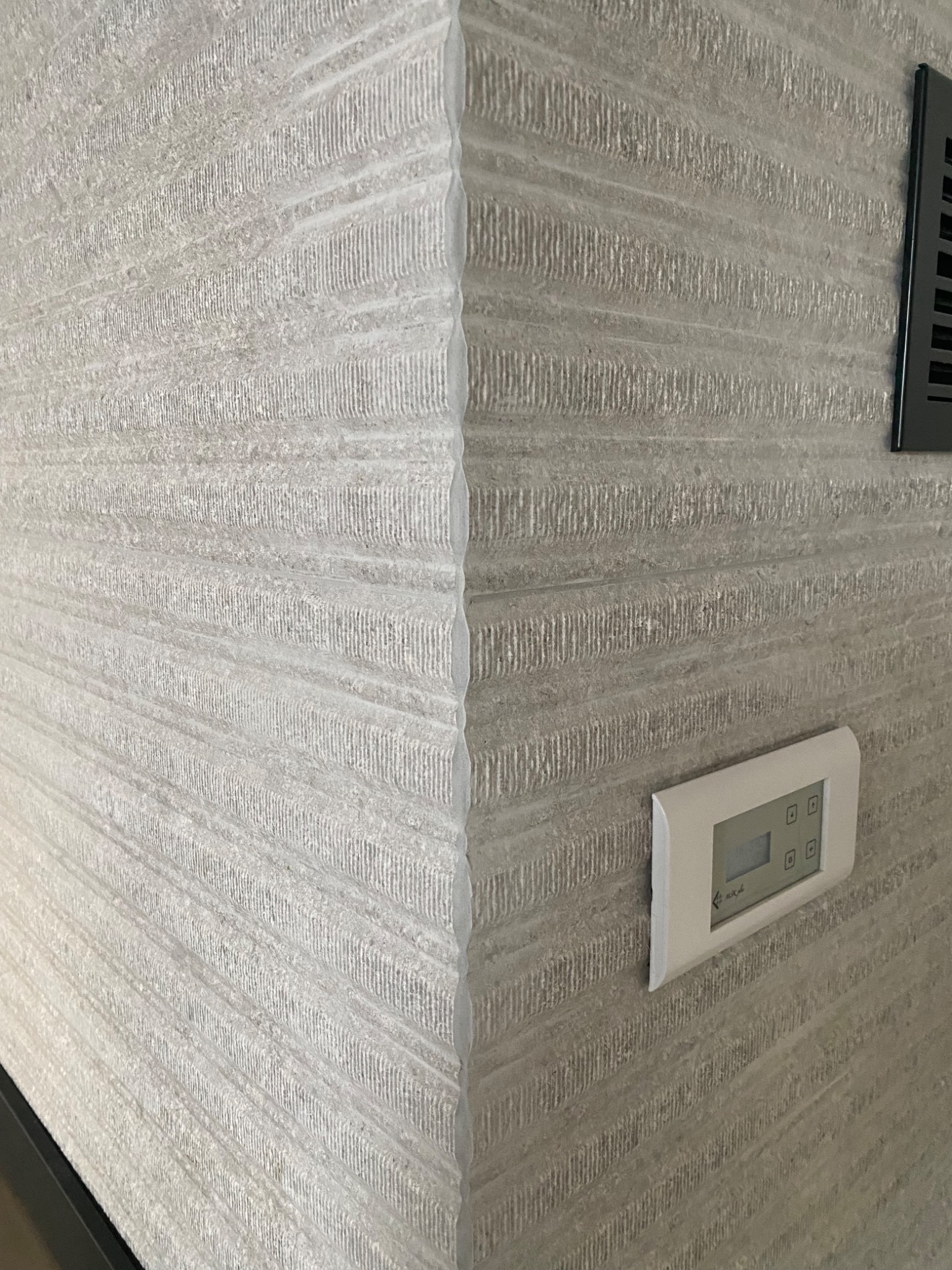 Szara ściana z poziomymi żłobieniami, widoczny narożnik, regulator temperatury i kratka wentylacyjna.