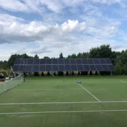 Instalacja fotowoltaiczna na trawiastym terenie, prawdopodobnie kort tenisowy, widoczne panele, rusztowanie i siatka tenisowa, w tle drzewa i pochmurne niebo.